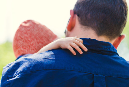 A child's hand is holding onto their father's shoulder, you see the back of the father's head with brown hair and the back of his dark blue shirt