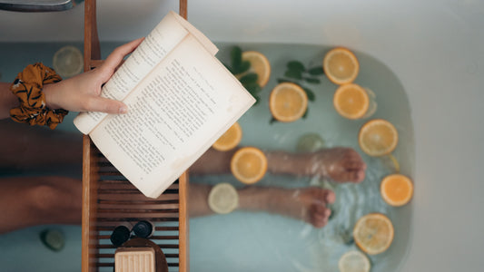 A birds-eye view of someone in the bath, the water is covered with herbs & citrus where you can see the person's legs. They are holding and reading a book.