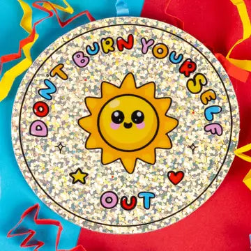 A round, glittery sticker featuring a smiling yellow sun in the center, surrounded by the text 'DON'T BURN YOURSELF OUT' in colorful letters, with small stars and hearts. The background is decorated with bright red, blue, and yellow confetti.