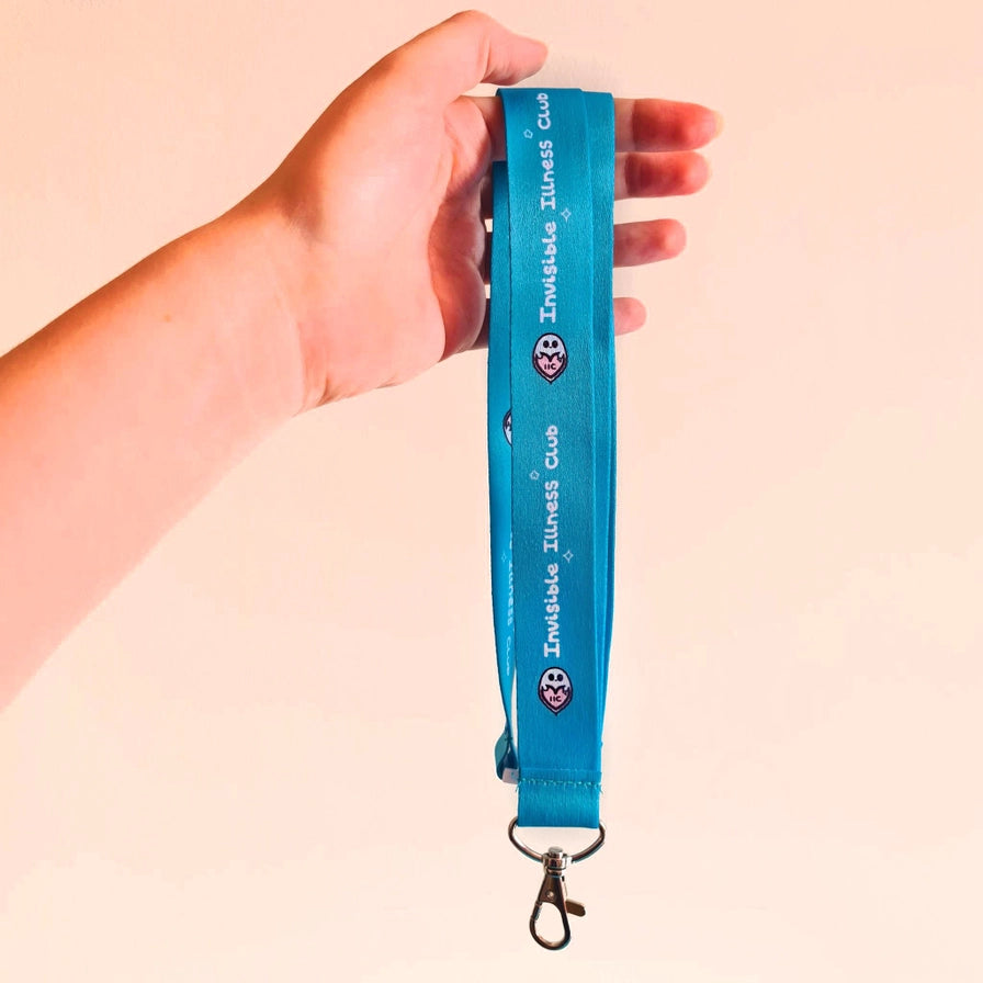A hand holding a bright blue lanyard strap with 'Invisible Illness Club' printed vertically in white, featuring small ghost logos, against a solid pale peach background.