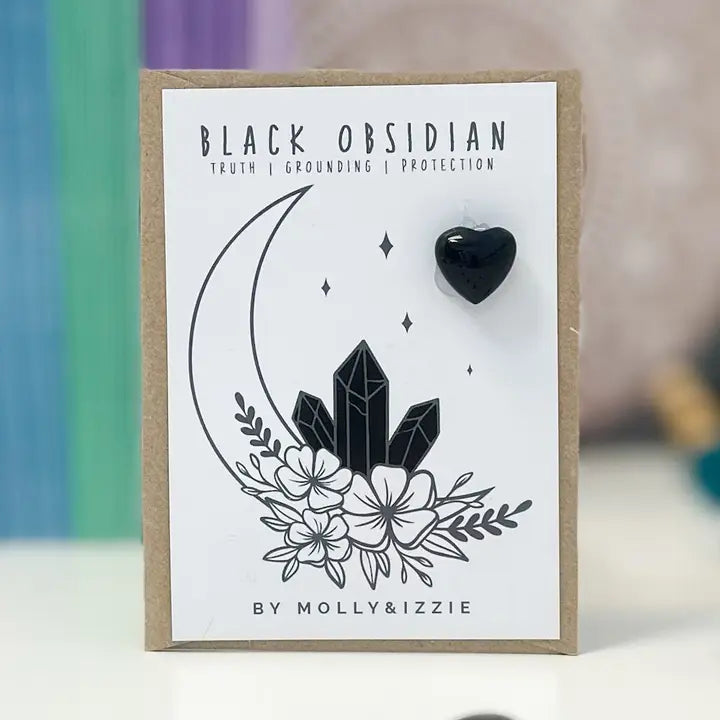A black obsidian heart-shaped crystal displayed on a paper backing card, which features a black crystal and moon illustrations with the text 'BLACK OBSIDIAN' and the brand 'BY MOLLY&IZZIE'.