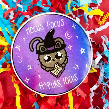 A round sticker with a purple and blue gradient background featuring a cartoon cat wearing a witch hat and holding a spellbook. The sticker is surrounded by colorful confetti. Text reads 'hocus pocus hypurr focus'.