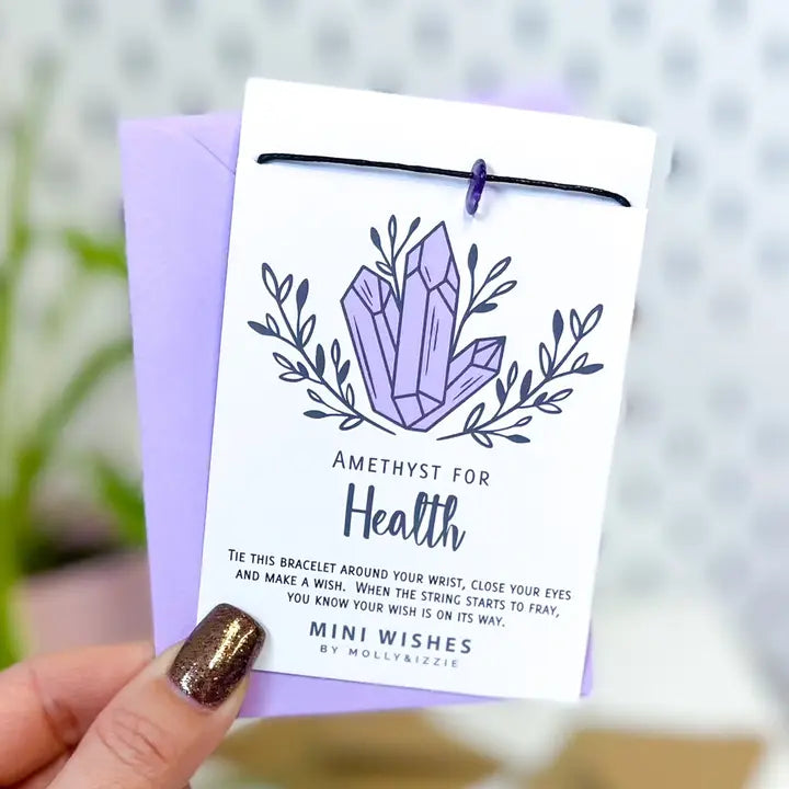 A hand with glittery brown nail polish holds a white card tied with a black string, showing a lavender amethyst crystal illustration and the text 'AMETHYST FOR Health'. A lavender envelope is visible behind the card, with a softly blurred light background.