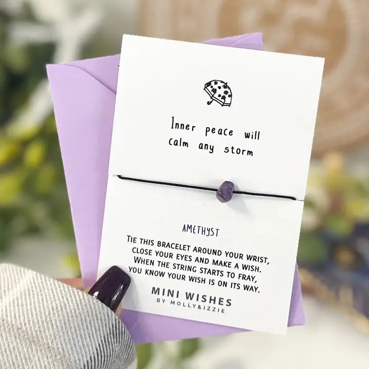 A white card with black text and a small umbrella illustration holds a thin black bracelet with a purple amethyst bead. A lavender envelope is behind the card, and a hand with dark nail polish is holding it. Text reads 'inner peach will calm any storm'.