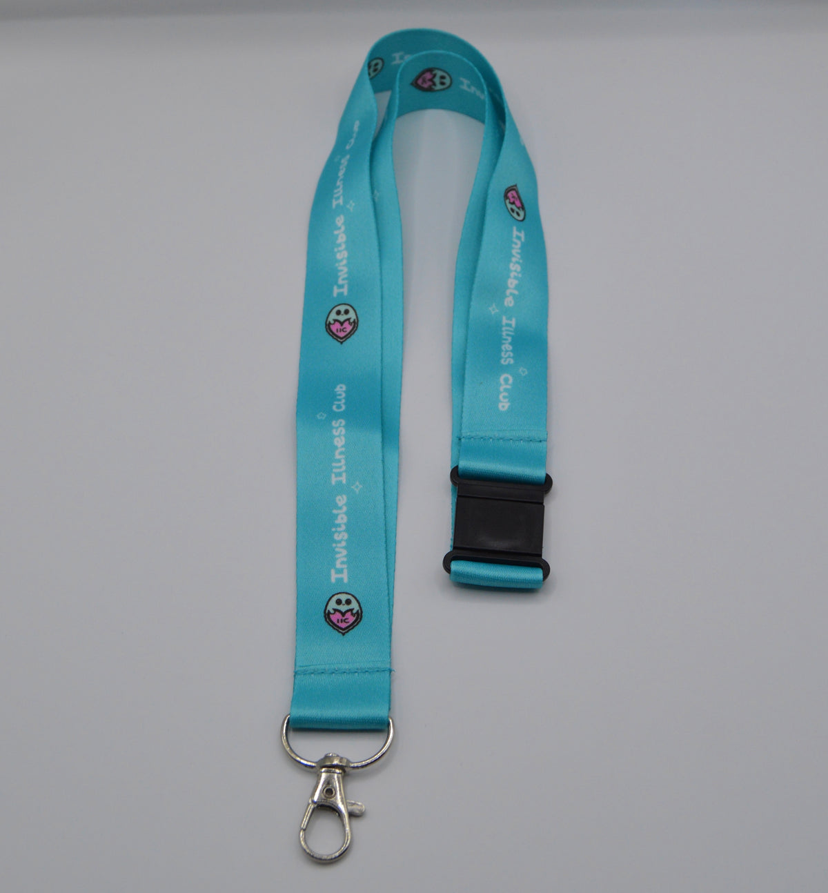 A turquoise blue lanyard with a black plastic quick-release buckle and a silver metal lobster clasp. The lanyard features white text reading 'Invisible Illness Club' and small ghost logos, laid flat on a neutral gray background.