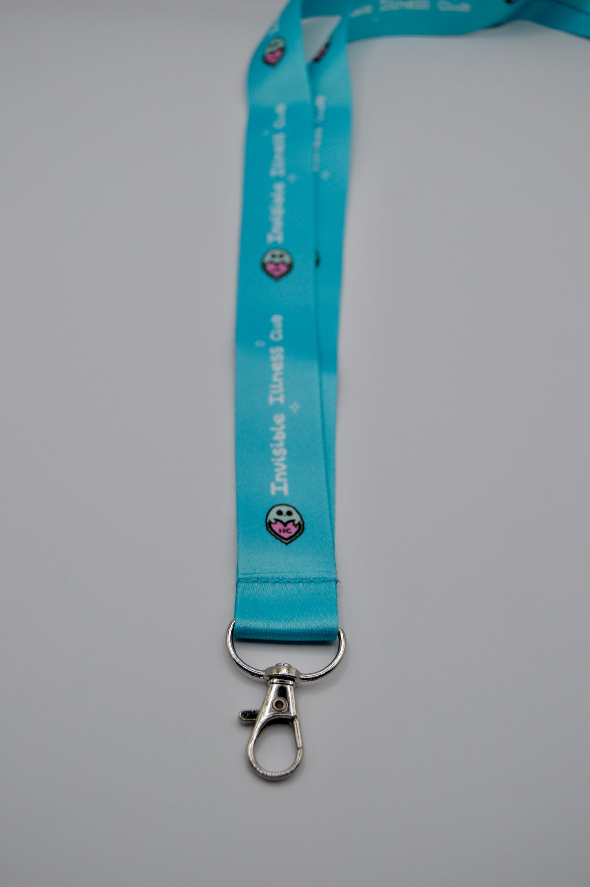 A bright turquoise blue lanyard strap with a silver lobster clasp, featuring the text 'Invisible Illness Club' and a small logo printed repeatedly along the strap, set against a plain light gray background.