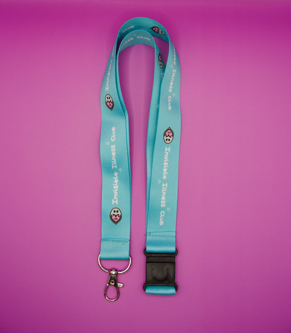 A turquoise blue lanyard with 'Invisible Illness Club' printed repeatedly in white, featuring small cartoon graphics and a black plastic quick-release buckle with a silver metal clasp, displayed on a solid pink background.