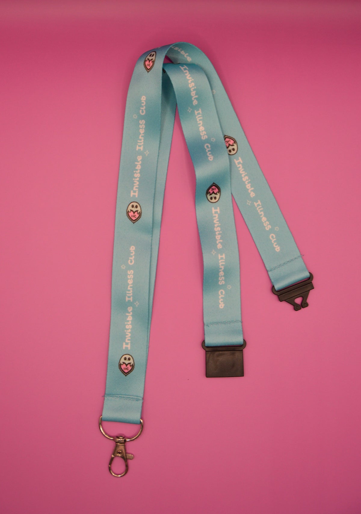 A light blue lanyard with 'Invisible Illness Club' printed repeatedly in white, featuring small pink and white ghost-heart graphics. The lanyard is laid flat on a vibrant pink background, with a silver metal lobster clasp on one end and a dark plastic buckle on the other.