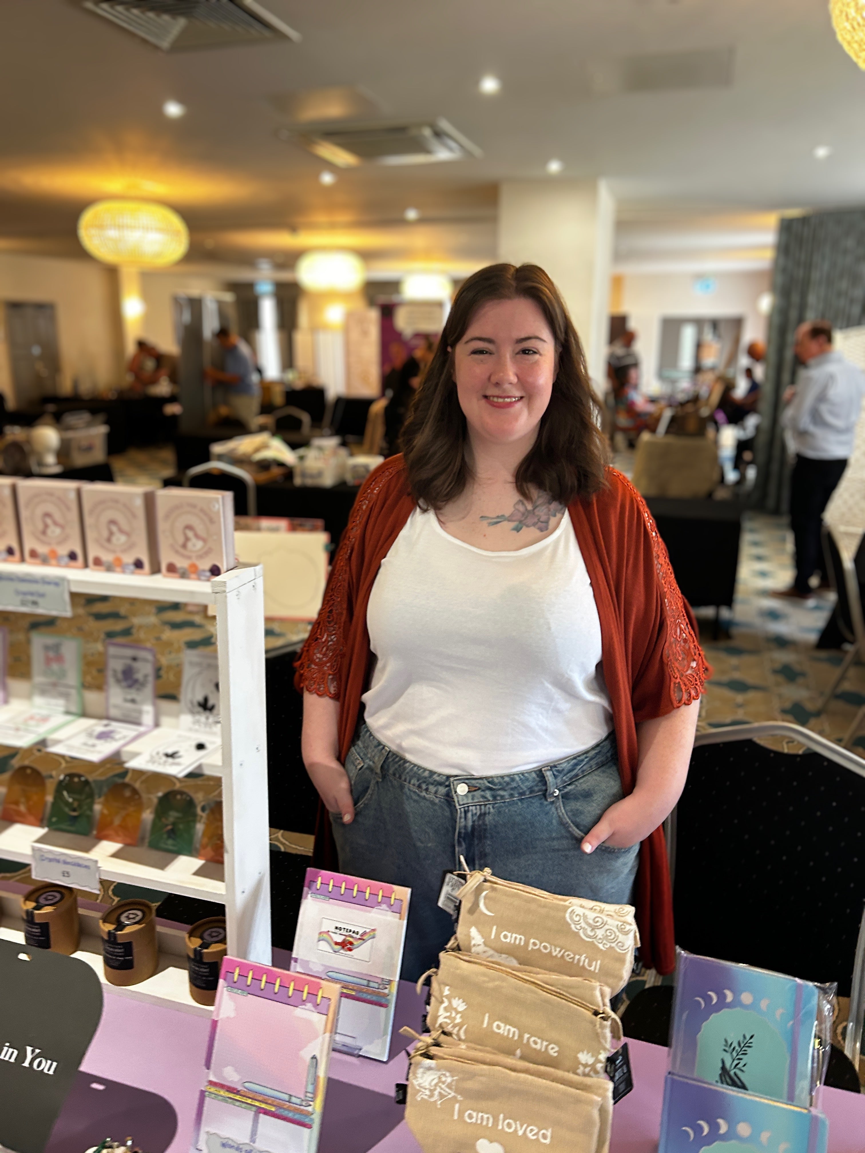 A smiling woman stands behind a display table in a busy indoor event space, wearing a white tank top, blue jeans, and a rust-orange cardigan. The table displays small packaged wellbeing items like journals and bath bombs.