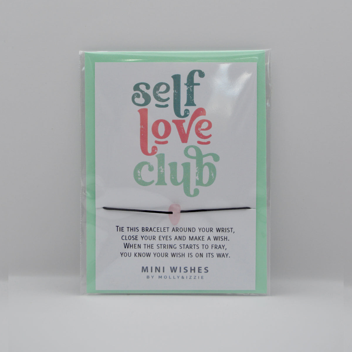 A packaged mini bracelet with a printed card that reads 'self love club' by Molly&izzie. The bracelet is a thin black cord with a small pink crystal bead, shown inside clear plastic packaging against a light gray background. Comes with a green envelope. 