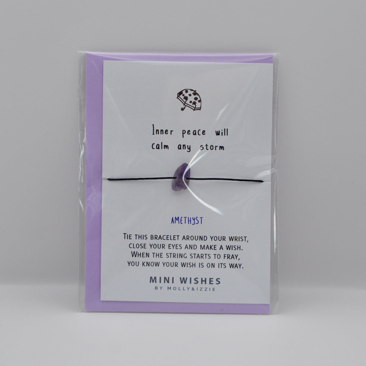 A purple amethyst crystal bracelet on a black string, presented on a lavender card with a clear plastic sleeve. The card features a small umbrella illustration and the text 'Inner peace will calm any storm'.