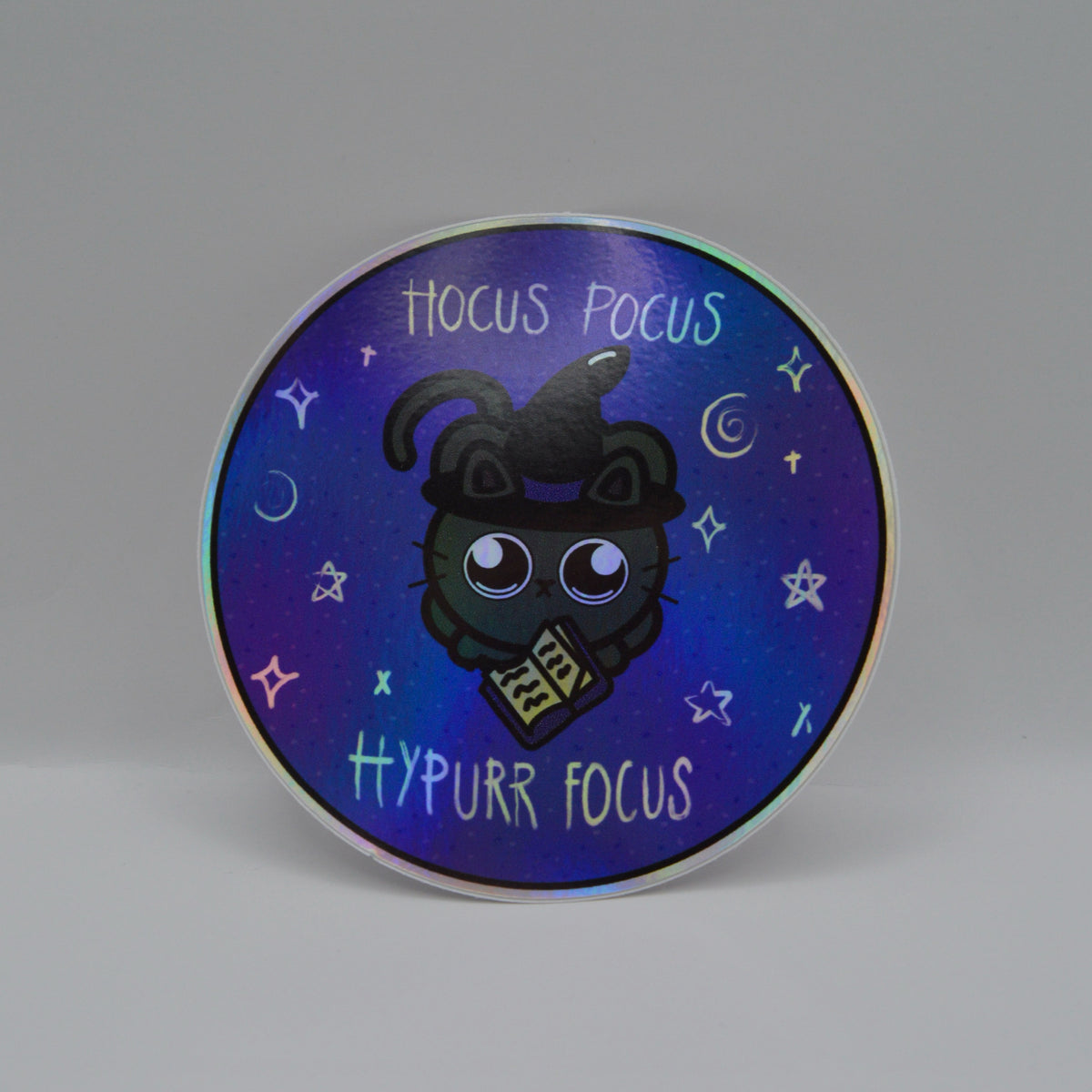 A round sticker with a purple and blue gradient background featuring a cartoon cat wearing a witch hat and holding a spellbook. Text reads 'hocus pocus hypurr focus'.