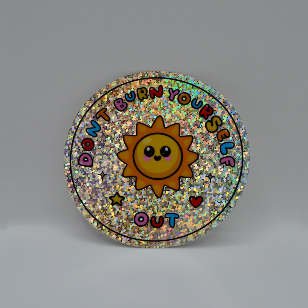 A round, glittery sticker featuring a smiling yellow sun in the center, surrounded by the text 'DON'T BURN YOURSELF OUT' in colorful letters, with small stars and hearts. 