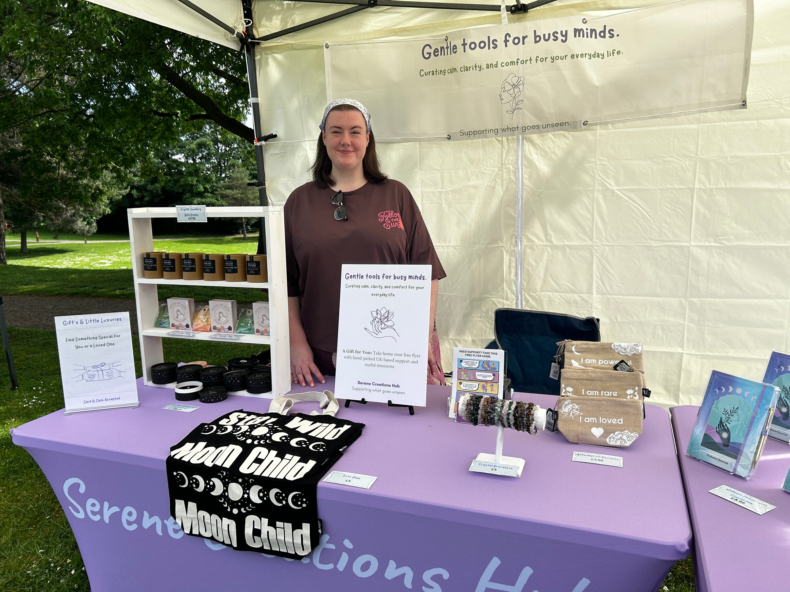 Person standing behind a table with products at an outdoor event, under a tent with branding.
