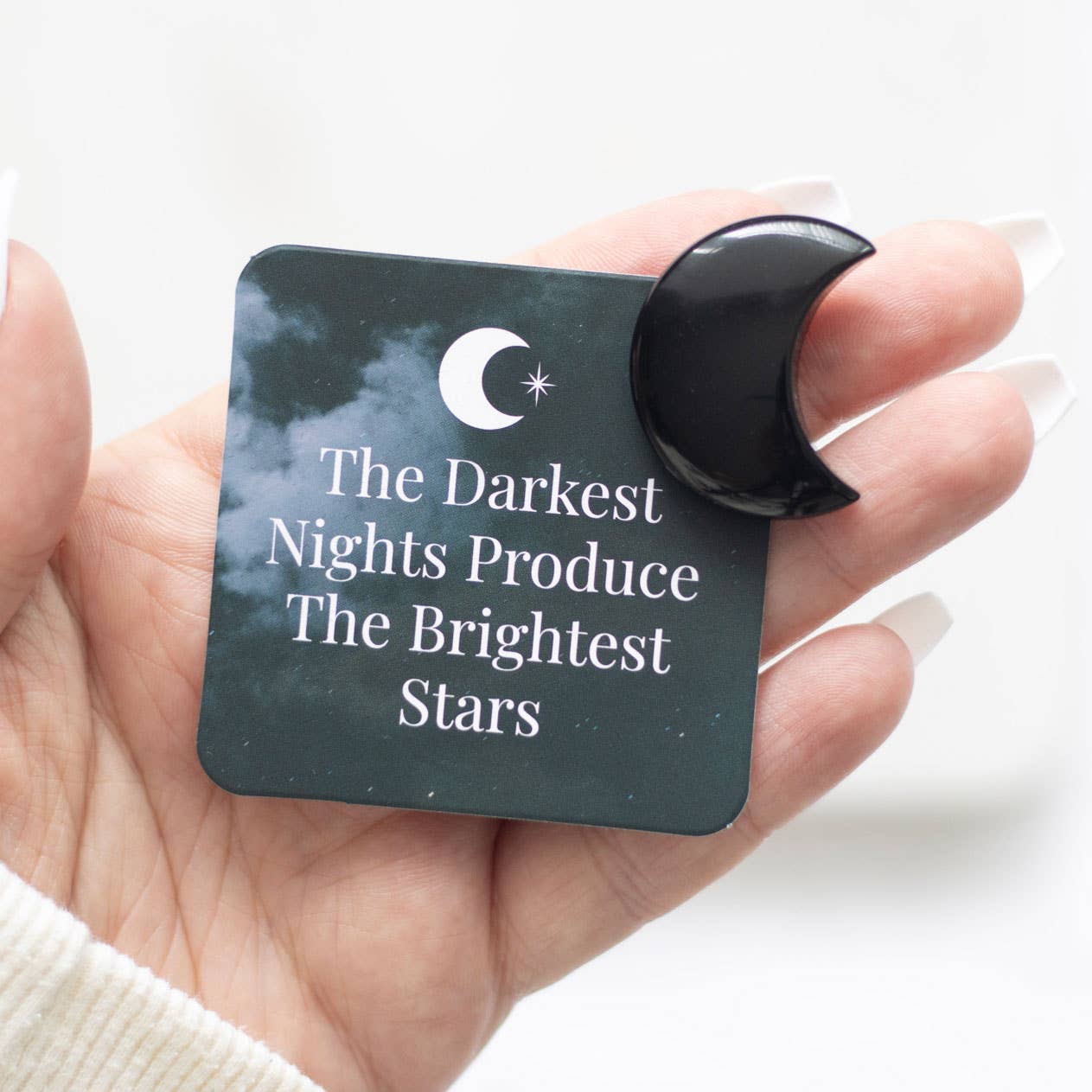 Hand holding a square card with a moon and star design and text 'The Darkest Nights Produce The Brightest Stars'.