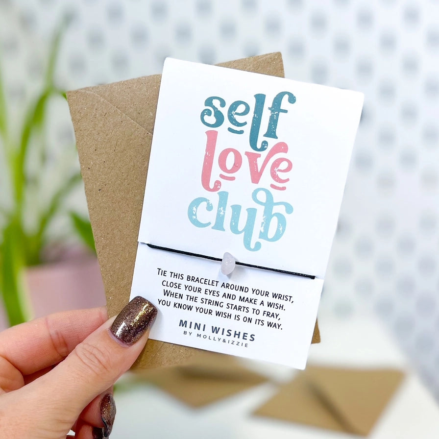 A hand with glittery nail polish holds a white card featuring a self-love bracelet, with a paper envelope behind it. The card reads 'self love club' in blue and pink lettering, with instructions and the brand 'bymolly&izzie' printed below.